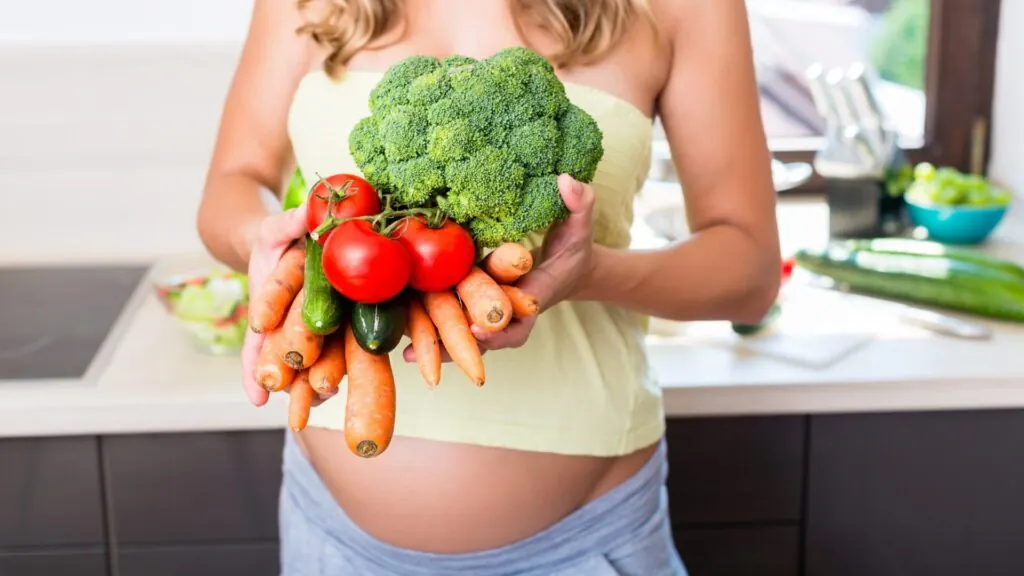 Gestante segurando brócolis, tomates e cenouras na cozinha