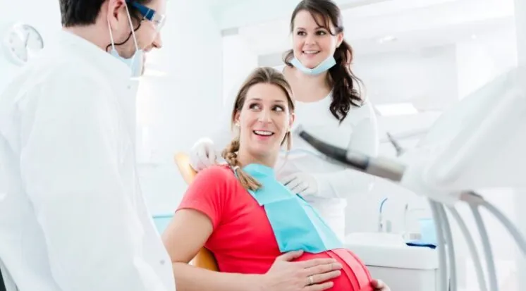Gestante sorrindo na cadeira odontológica, acompanhada pelo dentista e pela auxiliar