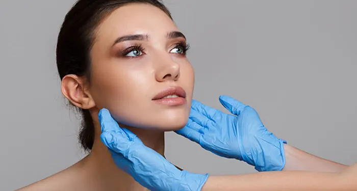 Mãos com luvas azuis apoiadas no rosto de uma mulher, como em uma avaliação facial