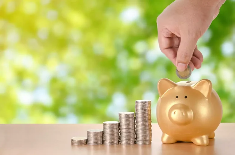 Mão colocando moeda em um cofrinho dourado em forma de porco, ao lado de pilhas de moedas